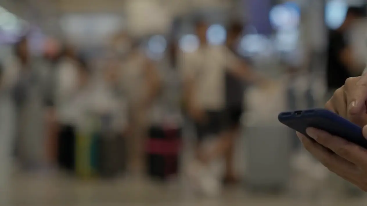Homem usando o celular no saguão de um aeroporto, com passageiros e bagagens desfocados ao fundo.
